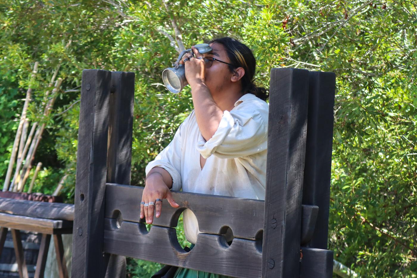 Person wearing glasses drinks from a metal cup while standing in wooden stocks outdoors, surrounded by leafy trees in a historical reenactment.