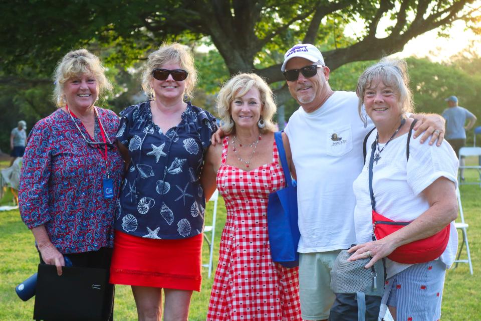 Five people smiling outdoors, standing in a park setting during sunset. They appear relaxed and happy, with trees and chairs in the background.