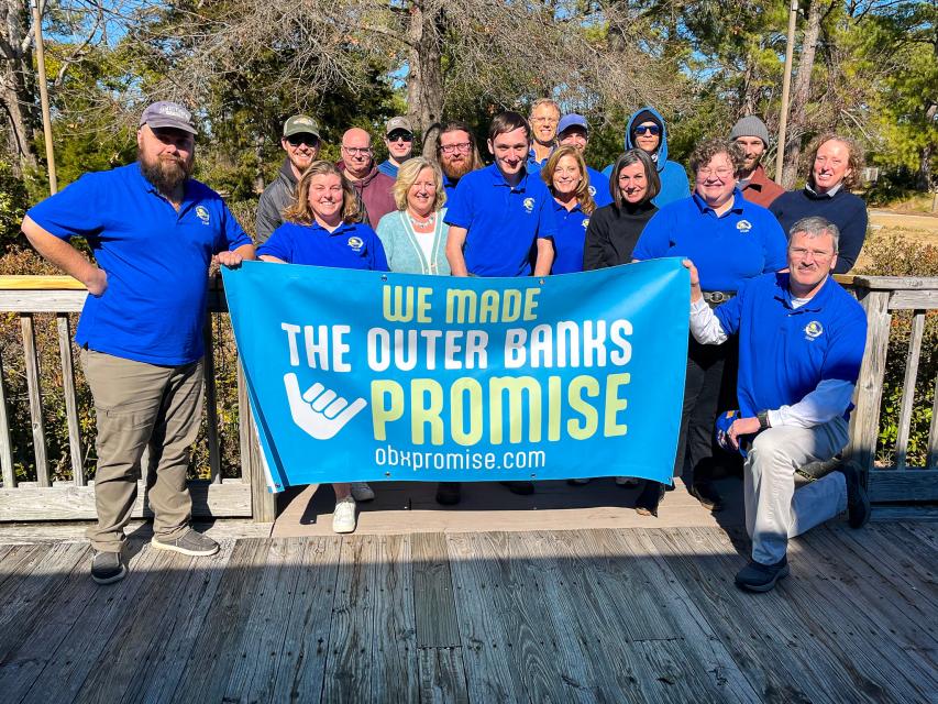 A cheerful group in blue shirts stands on a sunny wooden deck, holding an “Outer Banks Promise” banner with obpromise.com.