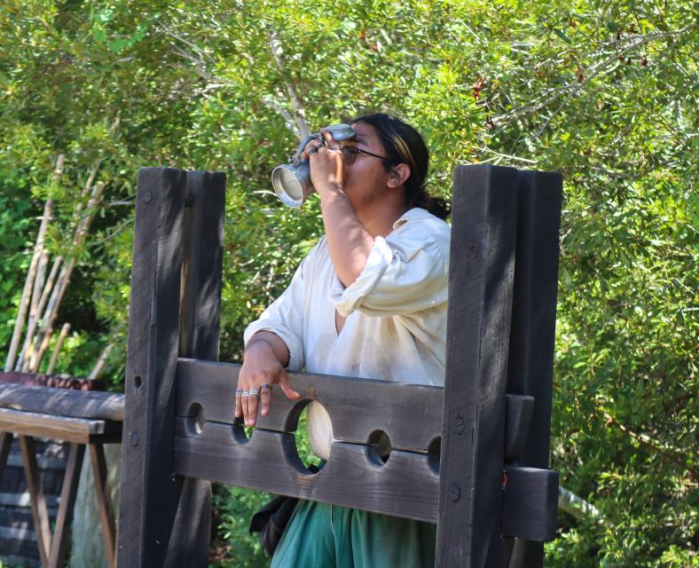 Person wearing glasses drinks from a metal cup while standing in wooden stocks outdoors, surrounded by leafy trees in a historical reenactment.