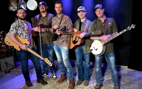 Picture of the band Monroeville. 5 people with different string instruments smiling at the camera from a stage. 