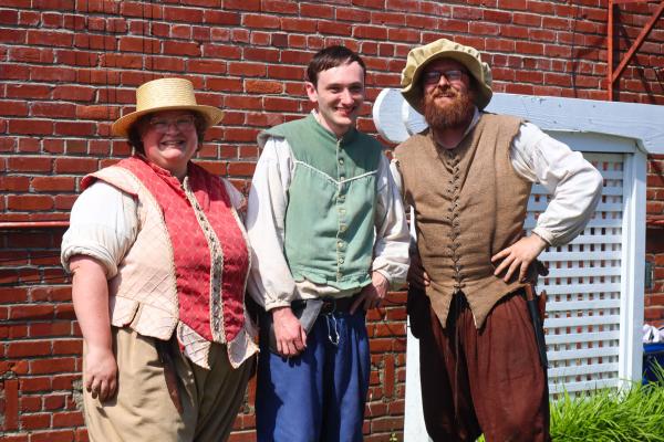 Three people dressed in historical costumes stand smiling in front of a brick wall. Their outfits are colorful, with vests and hats, conveying a cheerful, festive atmosphere.