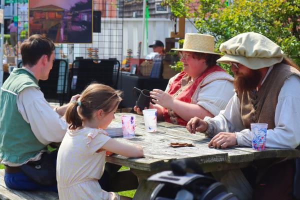 A family in historical costumes enjoys a sunny day at a picnic table, surrounded by art displays and greenery, conveying a cheerful vibe.