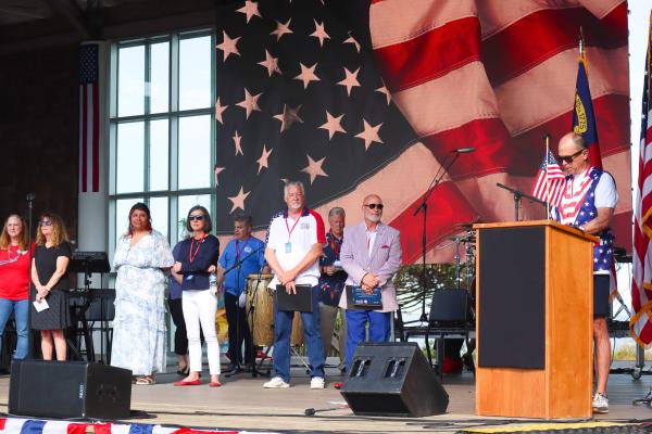 A group of people stands on a stage with a large American flag backdrop. A man at a podium speaks, holding a small flag. The atmosphere is patriotic.
