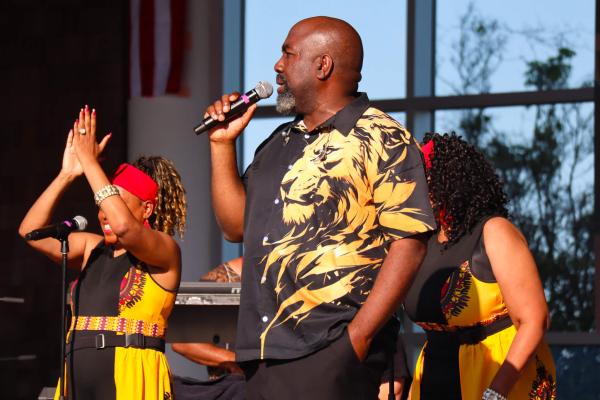 A man sings with two women on stage. He wears a black shirt with a lion design, while the women wear matching yellow dresses, creating a lively and energetic atmosphere.
