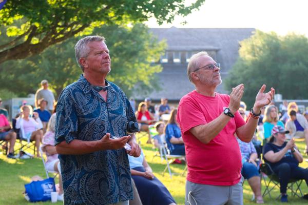 Two men standing outdoors; one wearing a blue patterned shirt, the other in a red shirt clapping. A seated audience is behind them with trees and a building in the background. The atmosphere is calm and engaged.