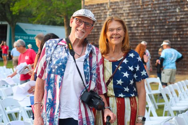 A smiling couple in American flag-themed outfits stand together outdoors, surrounded by white folding chairs. The mood is joyful and festive.