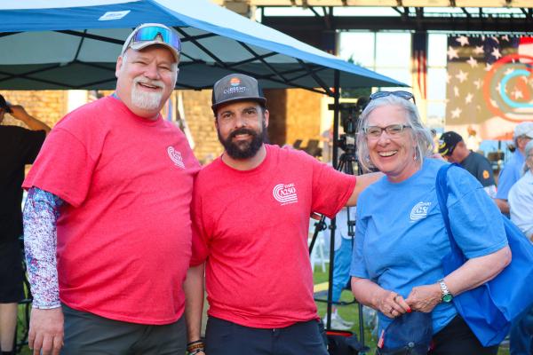 Three people smiling under a blue tent at an outdoor event. Two wear red shirts, one wears blue. The relaxed atmosphere has an American flag backdrop.