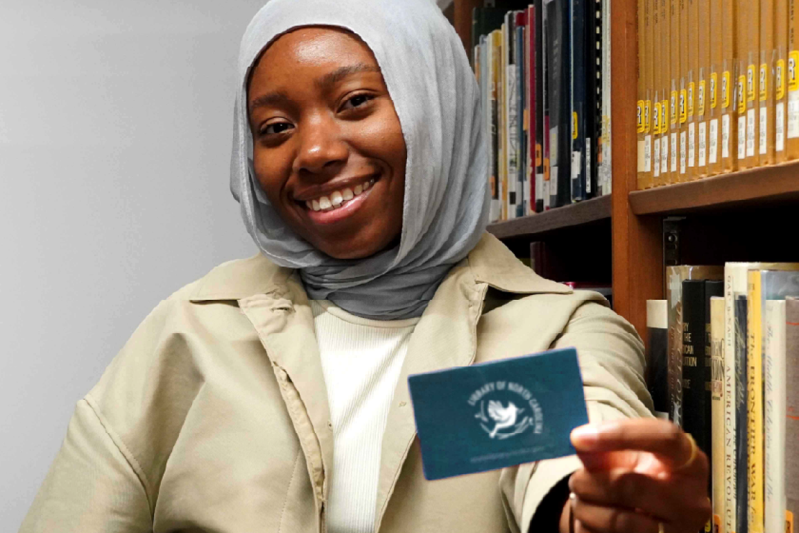 A Library of NC staff member happily holding her library card among the stacks
